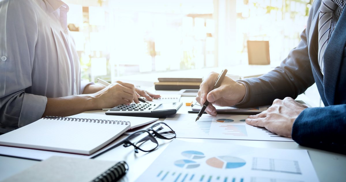 Duas pessoas conferindo relatórios financeiros com calculadora e caneta sobre a mesa